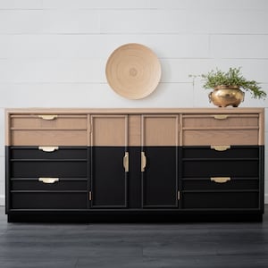 May include: A two-tone wooden dresser with a natural wood top and drawers, and black painted doors and drawers. The dresser has gold-colored hardware and is decorated with a wooden plate and a gold-colored pot with greenery.