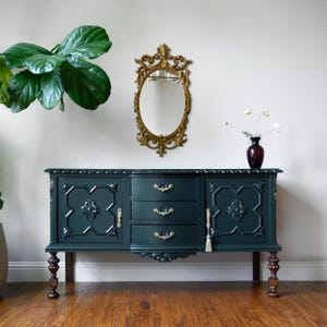 May include: A dark teal antique buffet with gold hardware and decorative accents. The buffet has three drawers and two cabinet doors. A gold oval mirror hangs above the buffet. A dark red vase with flowers sits on top.