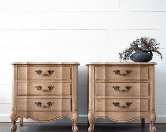 May include: Two matching wooden nightstands with three drawers each. The nightstands have a light brown finish, ornate bronze-colored handles, and curved legs. A dark vase with dried foliage sits atop one nightstand.