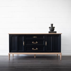 May include: A black sideboard with three drawers and two cabinet doors, featuring gold-colored hardware. The top is a light wood color, and the legs are also light wood. A dark vase and a book sit on top of the sideboard.