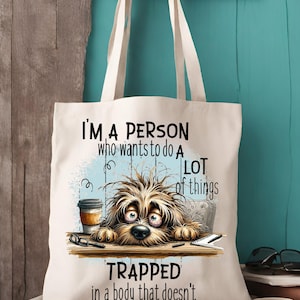 Pu&ograve; includere: Una borsa di tela bianca con un'illustrazione di un cane di cartone animato e il testo "Sono una persona che vuole fare molte cose intrappolata in un corpo che non lo fa".
