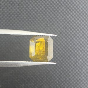Puede incluir: Una gema rectangular facetada de color amarillo dorado es sostenida por unas pinzas. El corte de la gema revela múltiples facetas, realzando su brillo. El fondo es una superficie texturizada de color gris oscuro. Medidas en milímetros.