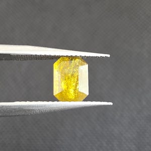 Puede incluir: Una gema amarilla facetada, de corte rectangular, es sostenida por unas pinzas sobre un fondo gris. La gema tiene un tono dorado brillante y parece transparente. Las pinzas son de color plateado y tienen un agarre texturizado.