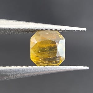 Puede incluir: Una gema facetada de color amarillo dorado sujeta con pinzas plateadas sobre un fondo gris oscuro. La gema tiene forma rectangular con múltiples facetas, que reflejan la luz. Las pinzas están enfocadas, resaltando la claridad y el color de la gema.