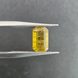 Puede incluir: Una gema amarilla tallada en forma rectangular, sujeta con pinzas. La gema tiene una superficie facetada e inclusiones internas. El fondo es gris neutro. Ideal para joyería.