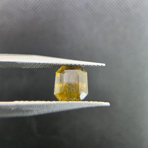 Puede incluir: Una gema facetada, de color amarillo verdoso, es sostenida por unas pinzas sobre un fondo oscuro. La gema tiene forma rectangular con esquinas cortadas y un aspecto ligeramente turbio, mostrando su belleza natural.