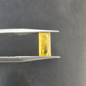 Puede incluir: Una gema rectangular amarilla es sostenida por unas pinzas plateadas sobre un fondo gris oscuro. La gema tiene una apariencia transparente y un tono dorado brillante. Las pinzas están bien enfocadas, resaltando la claridad de la gema.