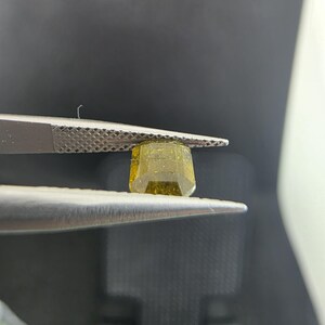 Puede incluir: Una gema facetada de color amarillo verdoso es sostenida por unas pinzas plateadas. La gema tiene forma rectangular con bordes biselados. Las pinzas están enfocadas, resaltando la claridad y el color de la gema.