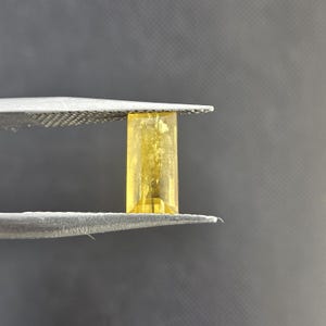 Puede incluir: Una gema rectangular, transparente y amarilla es sostenida por unas pinzas plateadas. La gema tiene una superficie pulida y lisa y un tono dorado vibrante. El fondo es gris oscuro.