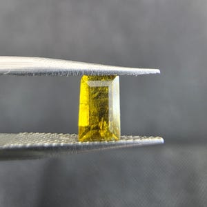 Puede incluir: Una gema rectangular amarilla es sostenida por unas pinzas plateadas. La gema tiene una apariencia ligeramente translúcida con estructuras internas visibles. El fondo es un gris oscuro borroso.