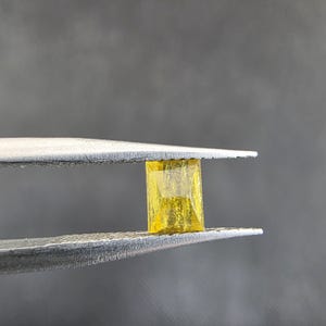 Puede incluir: Una gema rectangular amarilla es sostenida por unas pinzas plateadas sobre un fondo gris. La gema transparente tiene una superficie ligeramente texturizada, con estructuras internas visibles.