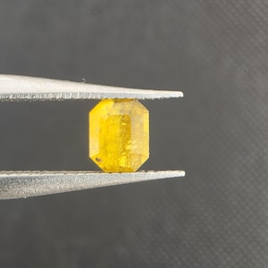 Puede incluir: Una gema amarilla tallada en forma rectangular es sostenida por unas pinzas. La gema tiene un aspecto brillante y translúcido con un tono dorado. Las pinzas son plateadas y el fondo es gris oscuro.