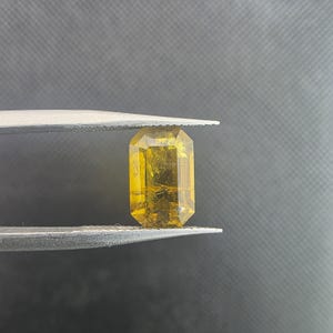 Puede incluir: Una gema amarilla tallada en forma rectangular es sostenida por unas pinzas sobre un fondo oscuro. La gema tiene un tono dorado con inclusiones internas y múltiples facetas.