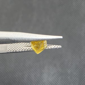 Puede incluir: Una pequeña gema amarilla facetada es sostenida por unas pinzas plateadas. La gema tiene una estructura cristalina y una apariencia translúcida. El fondo es de color gris oscuro sólido.