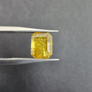 Puede incluir: Una radiante gema amarilla de corte rectangular sujeta con pinzas. La gema tiene un brillo dorado con texturas internas. Los bordes están cortados con precisión, reflejando la luz. El fondo es gris neutro.