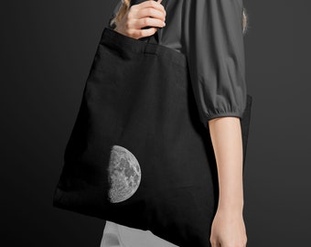 Op de afbeelding: Een zwarte tote bag met een witte halve maan print. De tas heeft lange hengsels en wordt door iemand gedragen.