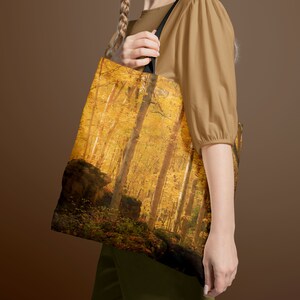 May include: Tote bag featuring a vibrant autumn forest scene. The image showcases tall trees with golden leaves, creating a warm, inviting atmosphere. The bag has black handles and is made of a durable material.
