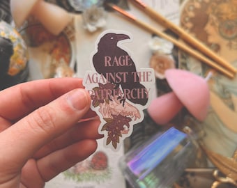 Może przedstawiać: Czarny kruk z czaszką i kwiatami siedzący na gałęzi. Kruk trzyma transparent z napisem "Rage Against The Patriarchy".