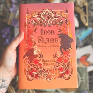 Może przedstawiać: Twarda oprawa książki zatytułowanej "Iron Flame" z serii Empyrean autorstwa Rebecci Yarros. Okładka przedstawia smoka, orła, elementy kwiatowe i celtyckie węzły na tle gradientu pomarańczowych i różowych odcieni.
