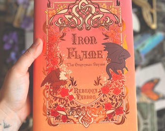 May include: A hardcover book titled "Iron Flame" from The Empyrean Series by Rebecca Yarros. The cover features a vibrant design with a dragon, an eagle, floral elements, and Celtic knotwork against a gradient of orange and pink hues.