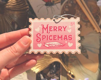 May include: A pink and white Christmas ornament in the shape of a postage stamp, with the words "MERRY SPICEMAS" and heart illustrations. The ornament is held in a hand, with a background of vintage ephemera and flowers.