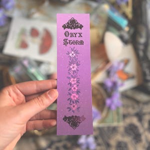 Op de afbeelding: Een paarse boekenlegger met de tekst "Onyx Storm" in een decoratief lettertype, versierd met bloemen en sierlijke ontwerpen. De boekenlegger heeft een gradiënteffect en wordt door een hand vastgehouden.