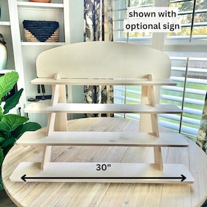 May include: A light-colored wooden display stand with four tiered shelves and a curved top panel. The base shelf features a black arrow graphic with the text 30 inches, and a text overlay in the upper right corner reads shown with optional sign.
