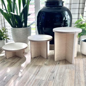 May include: Three wooden display stands of varying sizes, made of light-colored wood with a smooth finish. The stands are round and have four legs that form a cross shape.