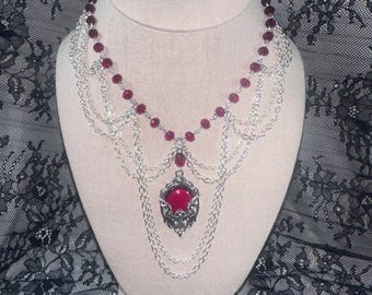 May include: A silver-toned necklace with a cascading design, featuring a large, oval, red gemstone pendant. The necklace has multiple strands of delicate chains and small, round, red beads. The necklace is displayed on a mannequin.