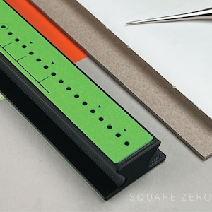 May include: A close-up of a bookbinding tool set. A black and green guide with evenly spaced holes is in the foreground. A needle with a wooden handle and a stack of paper are also visible.