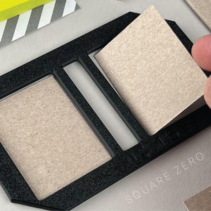 Peut inclure: Organisateur de bureau rectangulaire noir avec trois sections, contenant du papier de couleur beige. Une main retire une feuille de papier. Le texte "SQUARE ZERO" est visible. Un bloc-notes jaune est en arrière-plan.