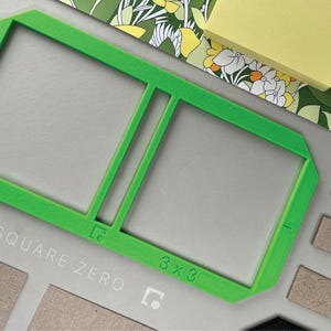 May include: A bright green 3x3 square frame with the text "SQUARE ZERO" and "3 x 3" printed on it. The frame is on a gray surface with other craft supplies, including a floral patterned paper and a stack of yellow sticky notes.
