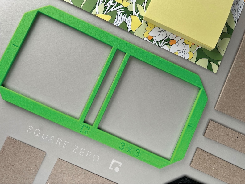 May include: A bright green 3x3 square frame with the text "SQUARE ZERO" and "3 x 3" printed on it. The frame is on a gray surface with other craft supplies, including a floral patterned paper and a stack of yellow sticky notes.
