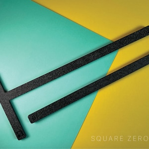 Op de afbeelding: Twee zwarte, getextureerde linialen op een turquoise en gele achtergrond. Eén liniaal is T-vormig, de andere is recht. De tekst "SQUARE ZERO" staat in witte letters in de rechter benedenhoek.