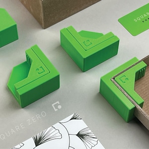 May include: Bright green corner clamps with the Square Zero Precision logo. The clamps are designed to hold cardboard pieces together. The image also shows a green business card and a box. The background is a light gray surface.