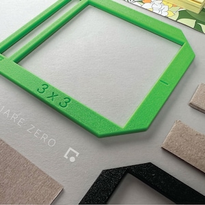 May include: A bright green 3x3 square frame with the text "SQUARE ZERO" in white. The frame is next to several rectangular pieces of cardboard and a black frame. A floral patterned notepad is in the background.