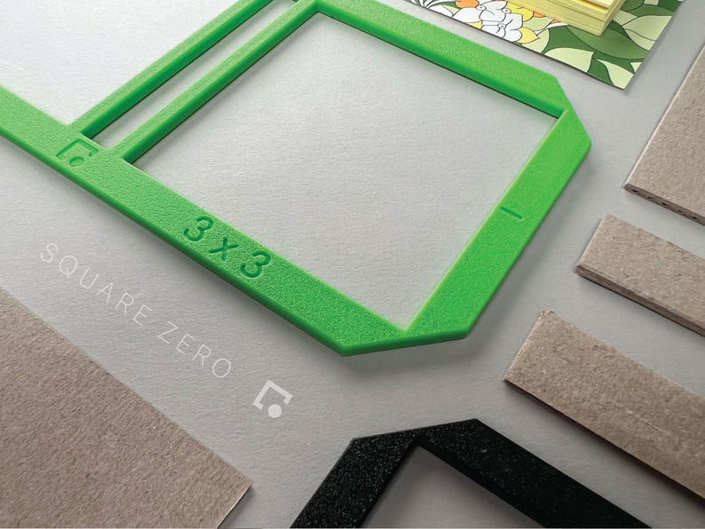 May include: A bright green 3x3 square frame with the text "SQUARE ZERO" in white. The frame is next to several rectangular pieces of cardboard and a black frame. A floral patterned notepad is in the background.