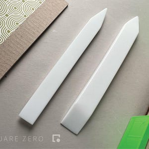 May include: Two white bone folders, angled, on a light grey surface. The tools are positioned near patterned paper and cardboard. The Square Zero logo is visible in the lower left corner.