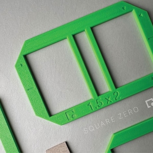 May include: Bright green plastic frame with two rectangular openings, labeled "1.5 x 2". The text "SQUARE ZERO" is printed below the frame. Additional green pieces are visible.