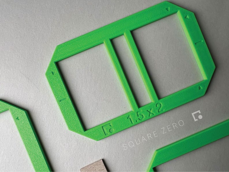 May include: Bright green plastic frame with two rectangular openings, labeled "1.5 x 2". The text "SQUARE ZERO" is printed below the frame. Additional green pieces are visible.