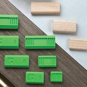 May include: An assortment of green and wooden dowels arranged on a wooden surface. The green dowels are rectangular with a series of vertical lines and numbers. The wooden dowels are rectangular with a textured surface. The text "SQUARE ZERO" is visible.