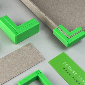 May include: Bright green L-shaped corner tools are positioned around a cardboard box. The tools are designed to fit the corners of the box. A green card with the text "SQUARE ZERO PRECISION" is also present.