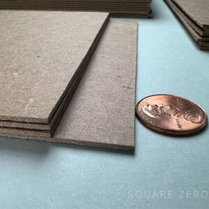 May include: Stacks of gray, rectangular cardboard sheets, possibly for crafting or packaging, are shown. A copper penny is next to the cardboard for size comparison. The image is on a light blue surface. The text "SQUARE ZERO" is at the bottom.