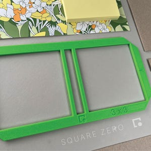 May include: A bright green 3x3 inch Square Zero frame, with a rectangular shape and a central divider. The frame is set against a light gray surface, with a floral patterned paper and sticky notes in the background.