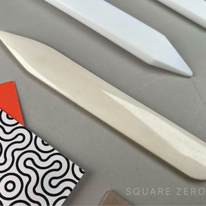 May include: Ivory bone folder tool with a rounded end and a pointed tip, used for paper crafting. Two white bone folders are in the background. A patterned paper and orange triangle are in the foreground. The Square Zero logo is in the bottom right corner.