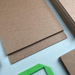 May include: Close-up of several stacks of brown cardboard sheets, with a green angled tool and a black strip of material. The text "SQUARE ZERO" is printed in light gray on the light blue surface.