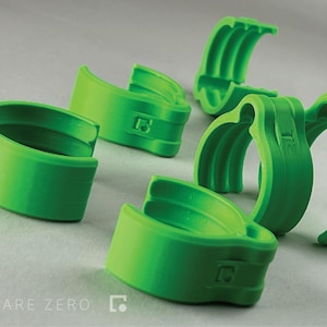 May include: Bright green plastic clips with a curved design. The clips have a smooth finish and feature the Square Zero logo. The image shows several clips arranged on a light gray surface.