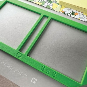 May include: Bright green 3x3 square frame with the text "SQUARE ZERO" and a logo. The frame is on a gray surface, with a floral patterned paper and a stack of yellow sticky notes in the background.