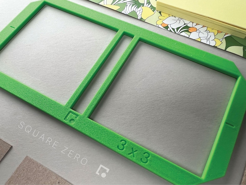 May include: Bright green 3x3 square frame with the text "SQUARE ZERO" and a logo. The frame is on a gray surface, with a floral patterned paper and a stack of yellow sticky notes in the background.