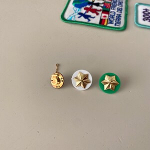 May include: Three gold-colored pins with a star design. The star pins are on a white and green background. The pin on the left has a number "5" on it. The pin in the middle is on a white background. The pin on the right is on a green background.